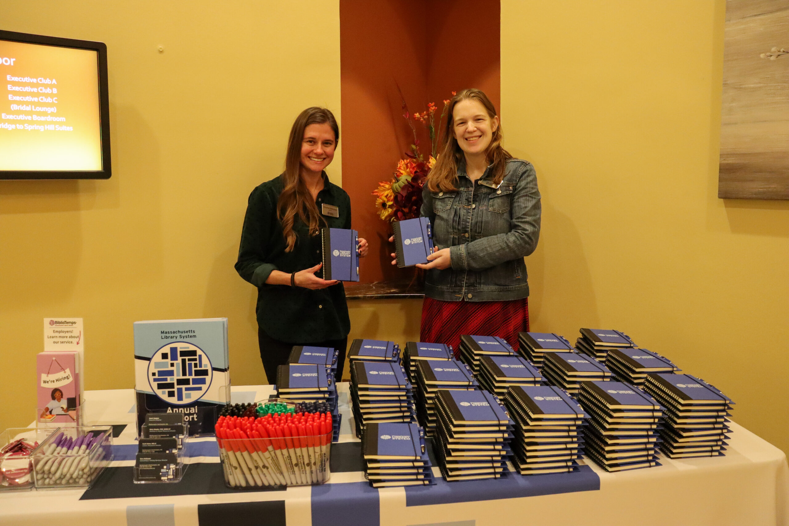 Two people holding MLS-branded notebooks in front of a table with MLS-branded promotional items.