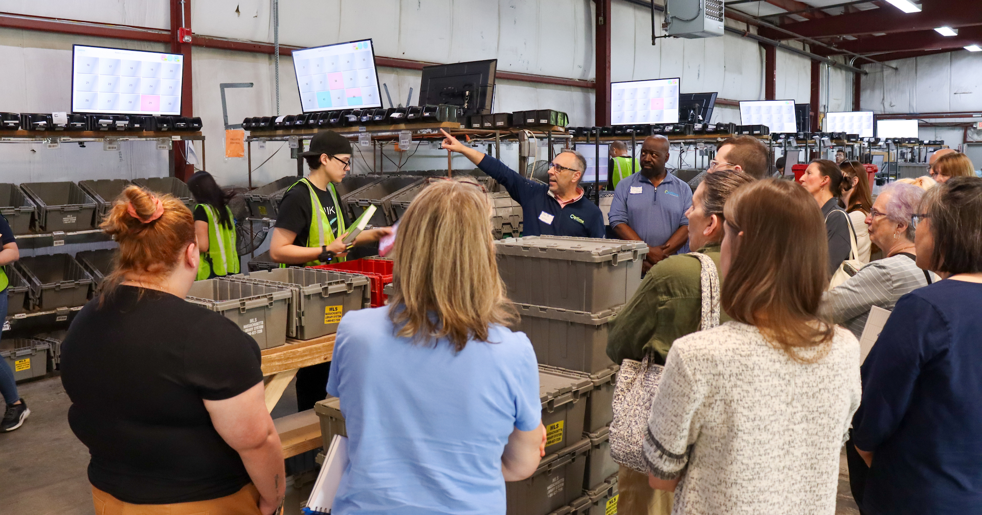 A group of a dozen people gathered around open sorting bins as an Optima Shipping employee points to informational screens.