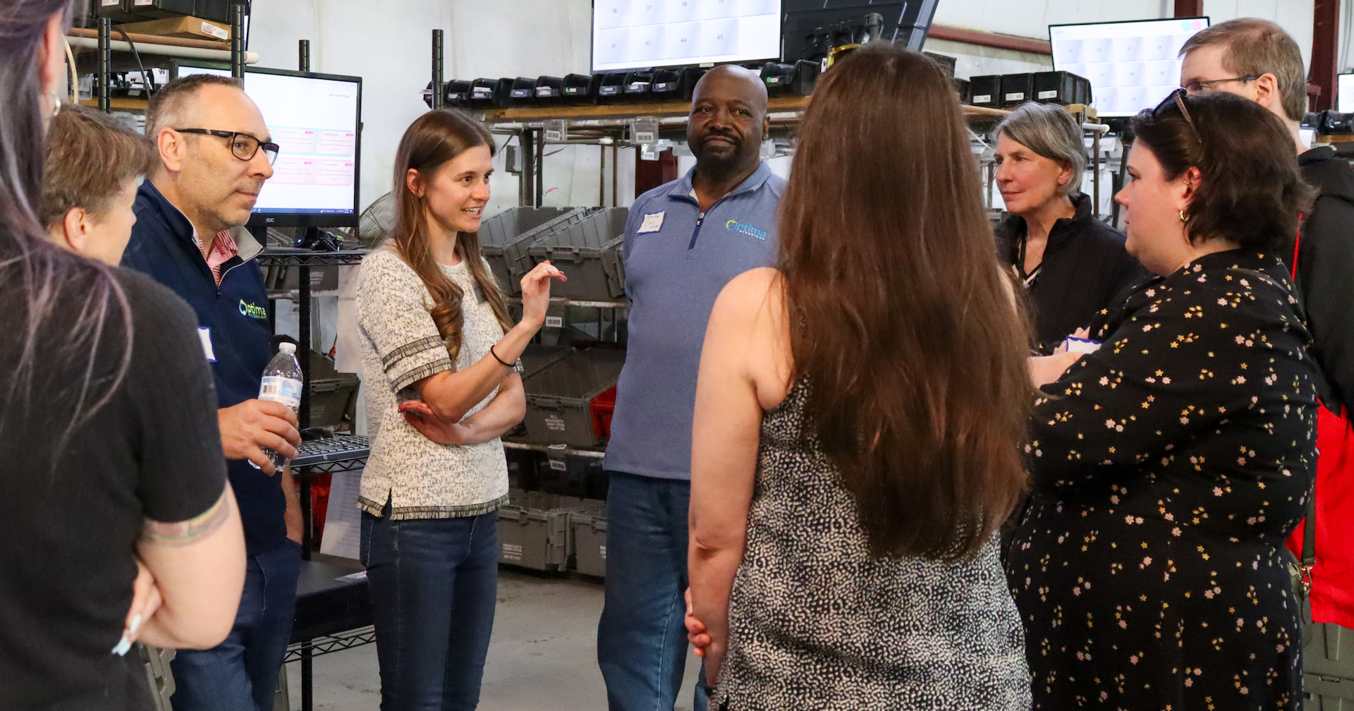 A group of people gather in a circle to listen to an MLS employee and two Optima Shipping employees.
