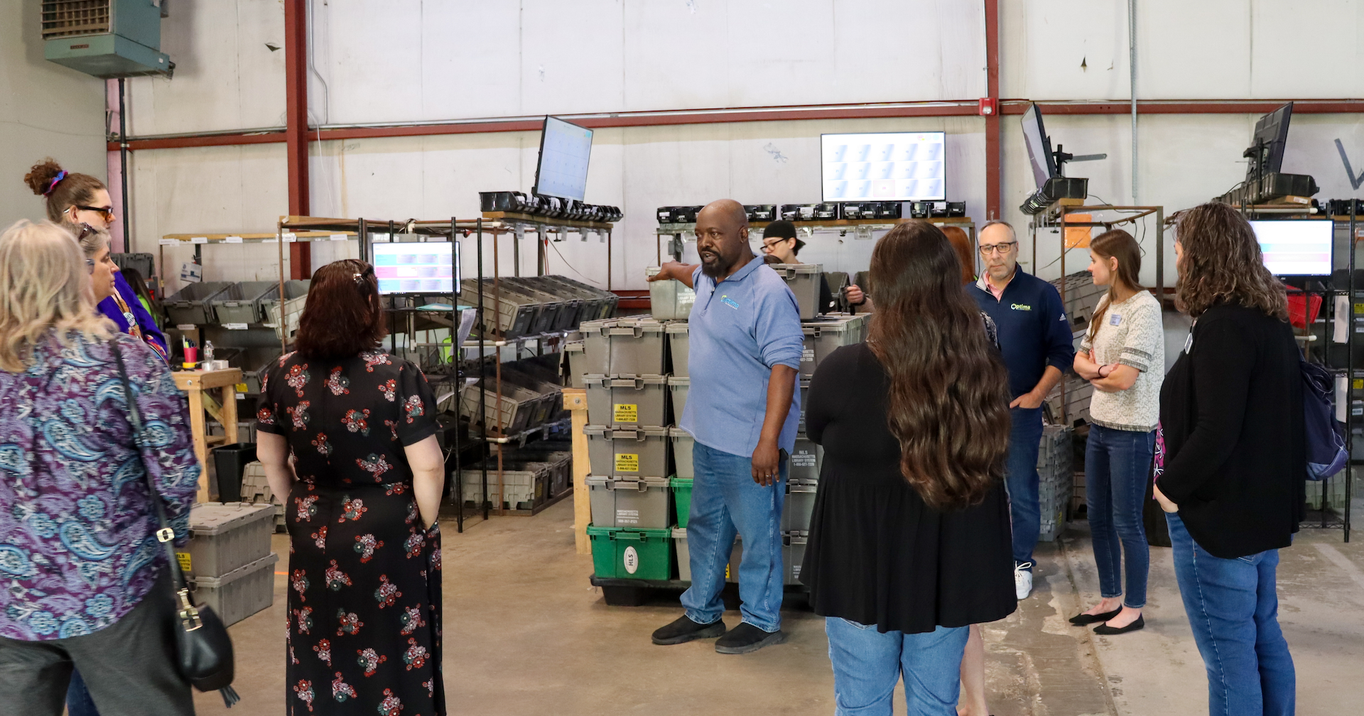 An Optima Shipping employee standing in front of stacks of sorting bins and speaking to a crowd of people.