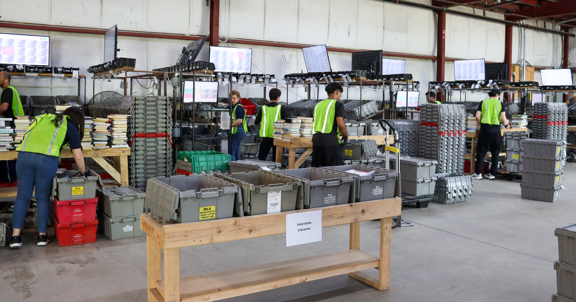 Half a dozen sorters in safety vests sort books in open sorting bins.